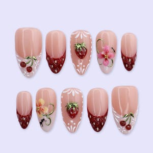 May include: A set of ten artificial nails with a light pink base and various designs. Some nails feature a red and white cherry design, while others have a strawberry or floral embellishment. The nails have a French tip design with a dark red pattern.