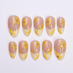 May include: A set of ten oval-shaped false nails. Each nail features a two-tone design with a light beige base and a nude pink upper section. Yellow floral designs and small gold accents adorn each nail, creating a delicate and feminine aesthetic.