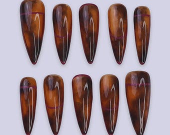 May include: A set of ten long, almond-shaped false nails. The nails have a gradient effect, transitioning from a dark brown at the tip to a lighter, amber hue. Each nail features a thin, horizontal purple stripe near the cuticle.