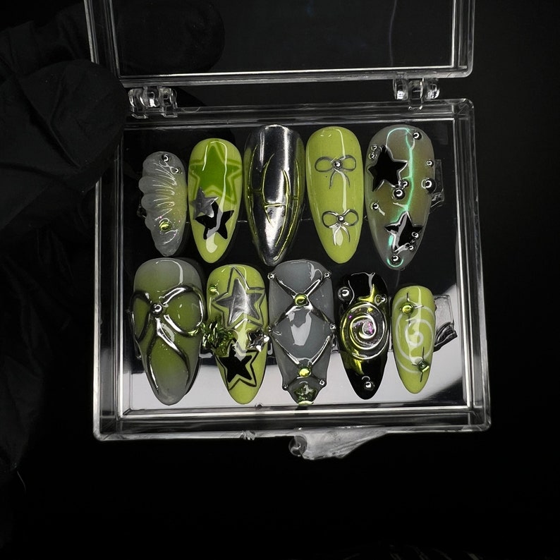 May include: A set of 10 green and black nail tips with various designs, including stars, bows, and swirls. The tips are displayed in a clear plastic case.