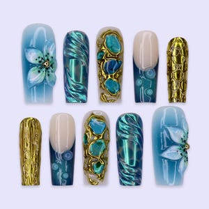 May include: A set of ten press-on nails featuring diverse designs including blue ombre with 3D white flowers, metallic teal swirls, gold-framed turquoise stones, dark blue French tips with dots, and textured gold foil patterns in various shapes.