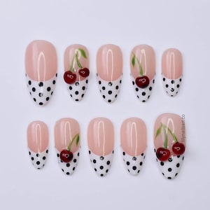 May include: Set of ten oval-shaped press-on nails. Each nail features a light pink base with a white polka dot design at the tip. A cherry design is painted on each nail. The nails are a glossy finish.