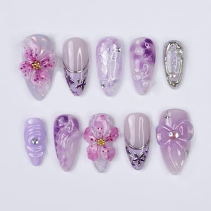 Pode incluir: Dez pontas de unhas roxas e brancas com vários desenhos, incluindo flores, borboletas, redemoinhos e laços. Os desenhos são pintados nas pontas e incluem glitter e pedras.
