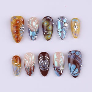 May include: A collection of ten artificial nails with various designs. The nails feature a range of colors, including orange, brown, blue, and yellow, with decorative elements like swirls, gems, and starbursts. Each nail has a unique pattern and shape.