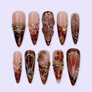 May include: A set of ten almond-shaped press-on nails featuring a nude base with brown leopard print patterns, gold metallic accents, raised floral designs, and small pearl embellishments. The nails are arranged in two rows of five on a light background.
