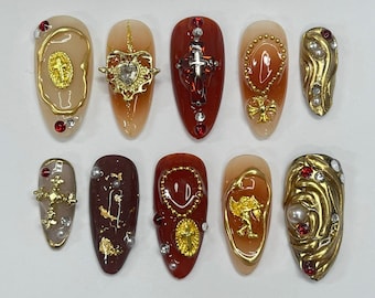 Op de afbeelding: Tien gouden nagelkunstontwerpen met verschillende religieuze en grillige motieven. De ontwerpen omvatten een kruis, een hart, een smiley en een kroon.