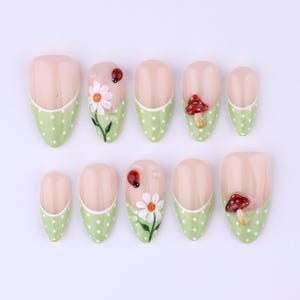Pode incluir: Conjunto de dez unhas artificiais com base bege clara e pontas verdes com bolinhas. Algumas unhas apresentam um desenho de joaninha, margarida branca ou cogumelo vermelho. As unhas são ovais.