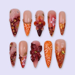 Puede incluir: Diez uñas artificiales con diversos diseños. Las uñas presentan una base nude con detalles intrincados, incluyendo adornos florales, acentos dorados y degradados de color en tonos naranja, rojo y marrón.