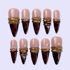 May include: A set of ten press-on nails with a pointed stiletto shape. Each nail features a nude base, a dark brown tortoiseshell tip, and a decorative band of gold foil, small white pearls, and clear rhinestones separating the two colors.
