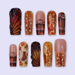 May include: A collection of ten false nails with diverse designs. The nails showcase autumnal patterns, such as butterfly wings, tartan, and leaf details. The colour palette includes brown, orange, and gold.