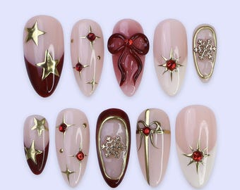 May include: A set of ten artificial nails with various designs. The nails feature a combination of light pink, burgundy, and gold accents. Some nails have gold stars, red gems, or a bow design. Others have a snowflake or bow design.