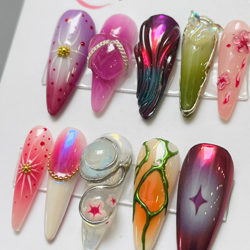 May include: Ten different nail art designs, each with a unique pattern and color scheme. The designs include a pink flower, a green leaf, a purple swirl, a white and pink flower, a white and gold star, a white and blue swirl, a white and green leaf, a white and orange swirl, a white and red flower, and a red and gold star.
