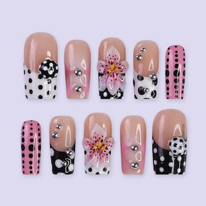 May include: A set of ten artificial nails with various designs. Some nails feature a nude base with black and white polka dots, while others have pink and black polka dots. Several nails are adorned with silver rhinestones and a 3D pink flower.