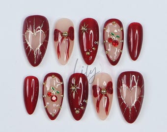 May include: Set of ten red and gold false nails with various designs. Some nails feature a heart, a bow, a cherry, and gold glitter.