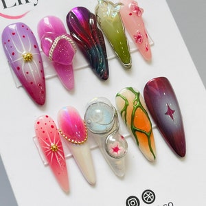 May include: Ten different nail designs featuring various colors, patterns, and textures. The designs include a pink floral design, a purple swirl design, a green and white abstract design, a pink and white geometric design, a clear design with a white star, a pink and white design with a gold accent, a clear design with a white and gold accent, a green and white design with a gold accent, a pink and white design with a gold accent, and a purple and white design with a gold accent.
