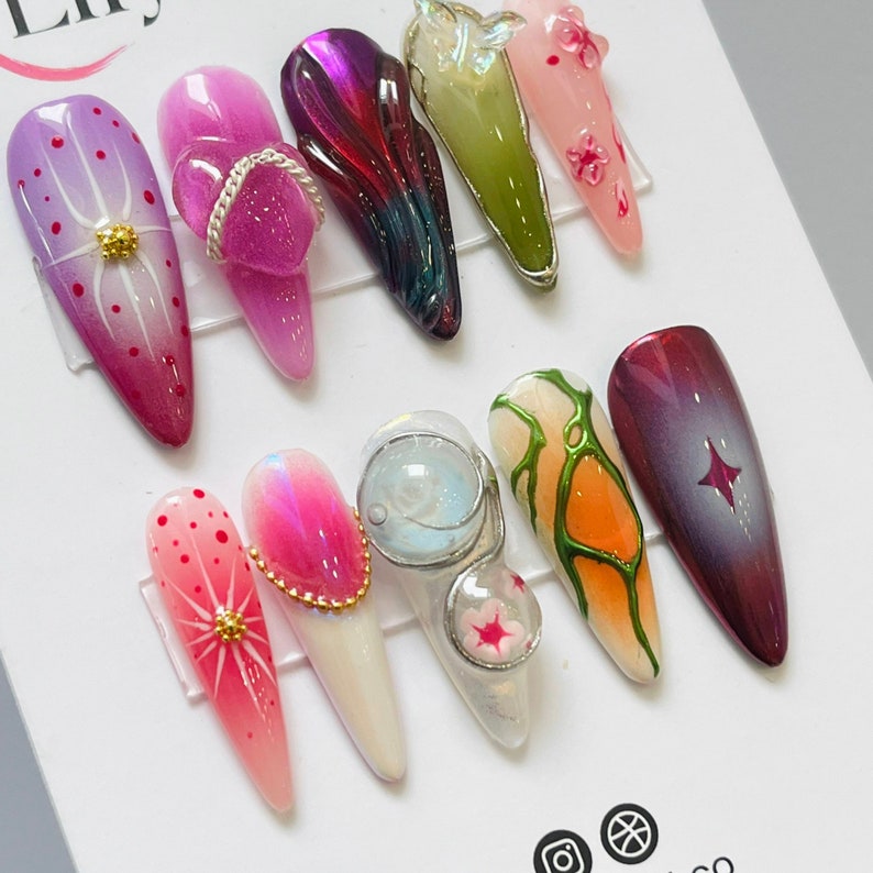 May include: Ten different nail designs featuring various colors, patterns, and textures. The designs include a pink floral design, a purple swirl design, a green and white abstract design, a pink and white geometric design, a clear design with a white star, a pink and white design with a gold accent, a clear design with a white and gold accent, a green and white design with a gold accent, a pink and white design with a gold accent, and a purple and white design with a gold accent.