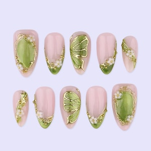 Könnte beinhalten: Set mit zehn künstlichen Nägeln in verschiedenen Designs. Die Nägel haben eine hellrosa Basis mit goldenen Akzenten, weißen floralen Details und grünen und goldenen Schmetterlings- und ovalen Designs. Die Nägel sind oval geformt.