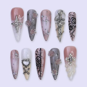 Pode incluir: Uma coleção de dez unhas artificiais longas e pontiagudas. Cada unha apresenta um design diferente, incluindo cruzes prateadas, rosas e adornos de espada. As cores incluem rosa, cinza e branco, com padrões intrincados e detalhes de pedras preciosas.
