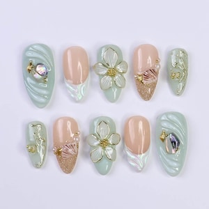 Puede incluir: Un conjunto de diez uñas artificiales con varios diseños. Algunas son de color verde claro con patrones ondulados y detalles iridiscentes. Otras son nude con purpurina, perlas y adornos de conchas marinas. Algunas presentan flores blancas y detalles dorados.
