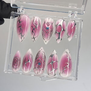 May include: A set of ten false nails in a clear plastic case. The nails are a pale pink color with a white ombre effect. Each nail has a different silver design, including swirls, hearts, and geometric shapes.