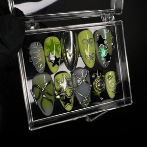 May include: A clear plastic box filled with 11 different nail art designs. The designs feature green, black, and silver colors with star and bow shapes.