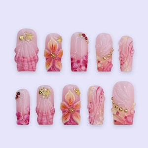 May include: A set of ten artificial nails with a light pink base. Each nail features intricate designs, including floral patterns, shell-like textures, and decorative gold and red accents. The nails are arranged on a light purple background.