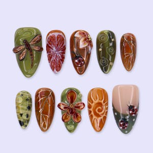 Puede incluir: Un juego de diez uñas postizas con formas almendradas y cuadradas con diseños inspirados en el otoño. La colección incluye motivos de una libélula, una flor, un sol, mariquitas y patrones abstractos en tonos verde oliva, naranja y marrón.