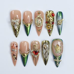 May include: A collection of ten almond-shaped artificial nails. Each nail has a nude base with detailed gold and jewel adornments. Designs include crosses, cherubs, hearts, and ornate frames, with metallic green tips.