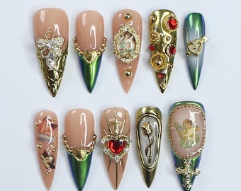May include: A set of ten almond-shaped artificial nails. Each nail features a nude base with intricate gold and jewel embellishments. Designs include crosses, cherubs, hearts, and ornate frames, with metallic green tips.
