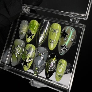 May include: A set of ten green and black nail art designs in a clear plastic case. The designs feature stars, bows, and swirls.