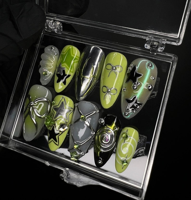 May include: A set of ten green and black nail art designs in a clear plastic case. The designs feature stars, bows, and swirls.