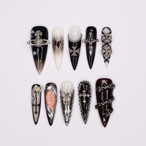Peut inclure: Dix ongles en forme de stiletto noirs et blancs avec divers ornements argentés et noirs. Les ornements comprennent un cœur, une croix, un papillon, un serpent et une chaîne.