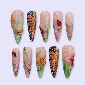 Puede incluir: Un conjunto de diez uñas artificiales con varios diseños. Las uñas presentan patrones de mariposas y flores en naranja, rosa y verde, con detalles dorados y una mariquita. Las uñas tienen forma de almendra y un acabado brillante.