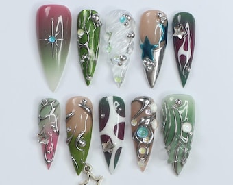 May include: Ten long, pointed, artificial fingernails with different designs. The designs include green and silver swirls, white and silver swirls, a teal star, a white and red design, and a green and silver design with a pink star.