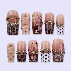 May include: A set of ten press-on nails featuring a nude base with various designs including brown leopard print, black polka dots, hand-painted brown floral motifs, and gold metallic accents with embedded clear rhinestones and small gold beads.