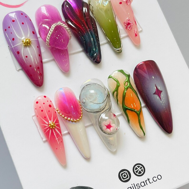 May include: Close-up of ten different nail designs. The designs include a variety of colors, patterns, and textures, including a white nail with a gold star, a pink nail with a gold chain, and a green nail with a white and orange design.