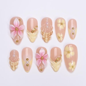 May include: A set of ten artificial nails in various designs. Each nail is a light pink color with decorative elements, including pink flowers, gold beads, and seashell accents. Some nails have yellow tips.
