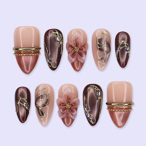 May include: A set of ten press-on nails featuring a mix of glossy nude and metallic brown finishes. Designs include three-dimensional floral accents, gold metallic line art, small embedded rhinestones, and horizontal gold bands with beaded textures.
