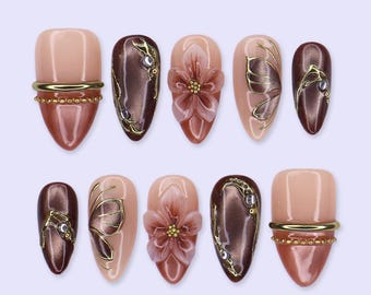 May include: A set of ten press-on nails featuring a mix of glossy nude and metallic brown finishes. Designs include three-dimensional floral accents, gold metallic line art, small embedded rhinestones, and horizontal gold bands with beaded textures.