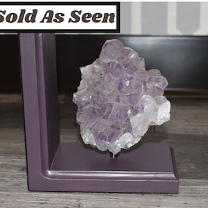 May include: A purple amethyst crystal cluster on a purple wooden stand. The crystal is clear and has a purple hue. The stand is a simple, rectangular shape.