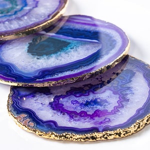 May include: Set of four purple agate coasters with gold trim. Each coaster features a unique pattern of purple and white agate.