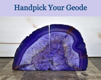 May include: A pair of purple agate geode bookends featuring concentric bands of deep violet and indigo hues surrounding a crystalline center. The text Handpick Your Geode is displayed in dark blue lettering above the two stone halves.