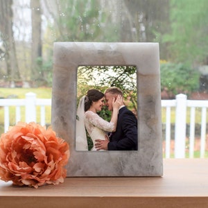May include: A white marble picture frame with a photo of a bride and groom kissing. The frame is sitting on a wooden surface with a peach-colored flower in the foreground.