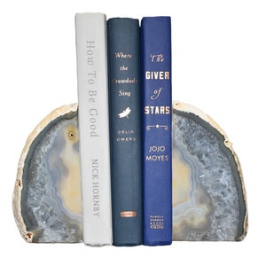 May include: Three hardcover books with white, blue, and dark blue covers are displayed on a gray and white agate bookend. The book titles are "How To Be Good" by Nick Hornby, "Where the Crawdads Sing" by Delia Owens, and "The Giver of Stars" by Jojo Moyes.