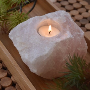 May include: A pink rose quartz candle holder with a small white tea light candle burning inside. The holder is on a wooden tray with a brown and white patterned surface.