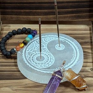 May include: A white selenite incense holder with a yin yang design and a mandala pattern. Two incense sticks are in the holder. A black lava rock bracelet with rainbow chakra beads is in the background. Two crystal pendants, one amethyst and one citrine, are in the foreground.