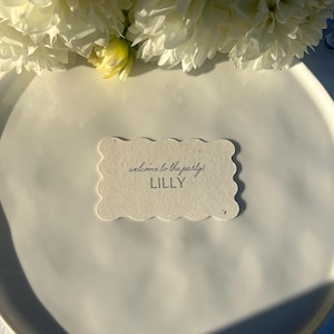 K&ouml;nnte beinhalten: Eine wei&szlig;e Tischkarte mit gewelltem Rand, auf der der Text "welcome to the party!" und der Name "LILLY" in schwarzer Schrift stehen, liegt auf einem wei&szlig;en Teller. Wei&szlig;e Blumen sind im Hintergrund.