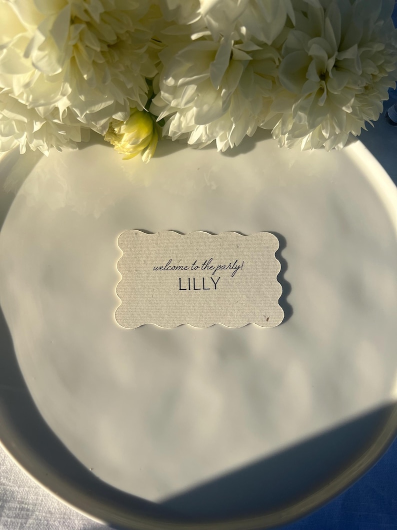 K&ouml;nnte beinhalten: Eine wei&szlig;e Tischkarte mit gewelltem Rand, auf der der Text "welcome to the party!" und der Name "LILLY" in schwarzer Schrift stehen, liegt auf einem wei&szlig;en Teller. Wei&szlig;e Blumen sind im Hintergrund.
