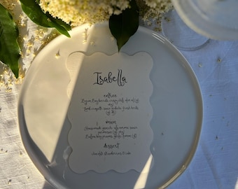 Könnte beinhalten: Ein weißer Teller mit einer Menükarte mit gewelltem Rand mit der Aufschrift "Isabella". Die Speisekarte listet eine Vorspeise, einen Hauptgang und ein Dessert auf. Der Teller steht auf einer weißen Tischdecke, mit Grün und einem Glas im Hintergrund.