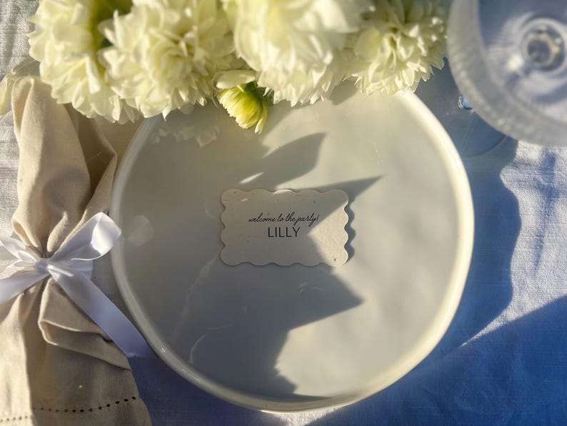 K&ouml;nnte beinhalten: Ein wei&szlig;er Keramikteller mit einer Namenskarte mit Wellenrand mit der Aufschrift "welcome to the party! LILLY". Der Teller ist mit einer wei&szlig;en Serviette mit wei&szlig;em Band und wei&szlig;en Blumen gedeckt. Im Hintergrund ist ein Weinglas zu sehen.